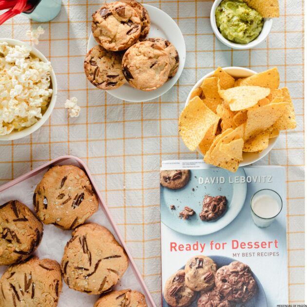 Una mesa con galletas, papitas, guacamole y un libro.