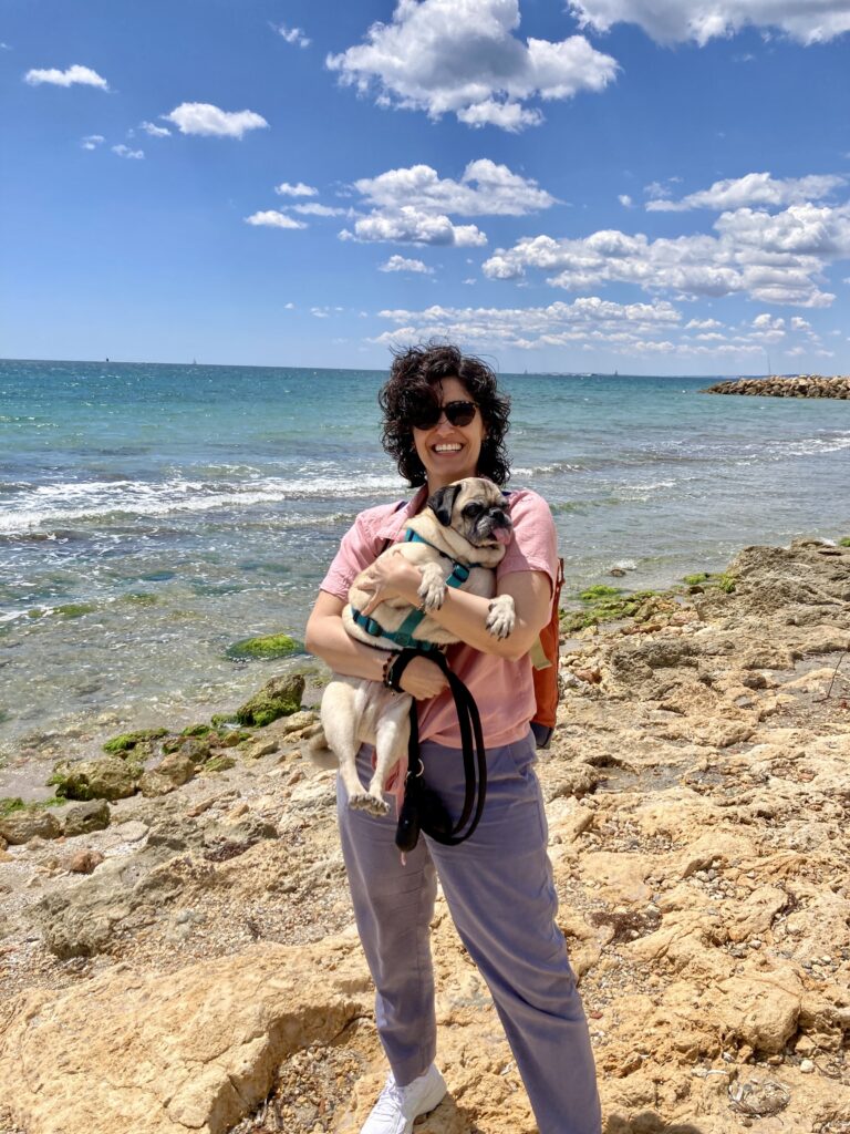 Una mujer sosteniendo un pug en una playa rocosa.