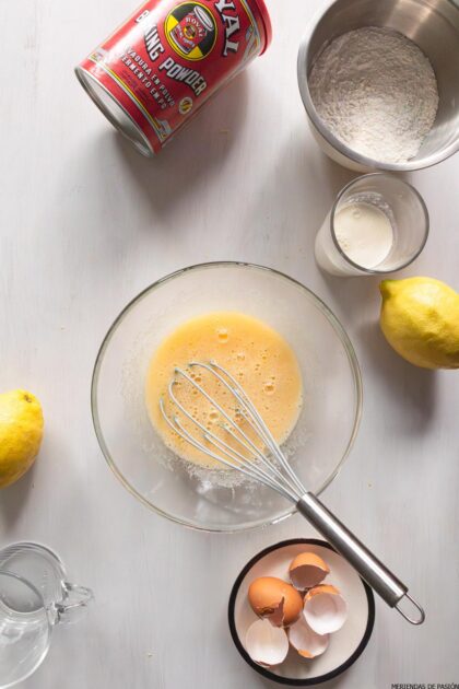 Un cuenco con huevos, limones y batidores sobre una mesa blanca.