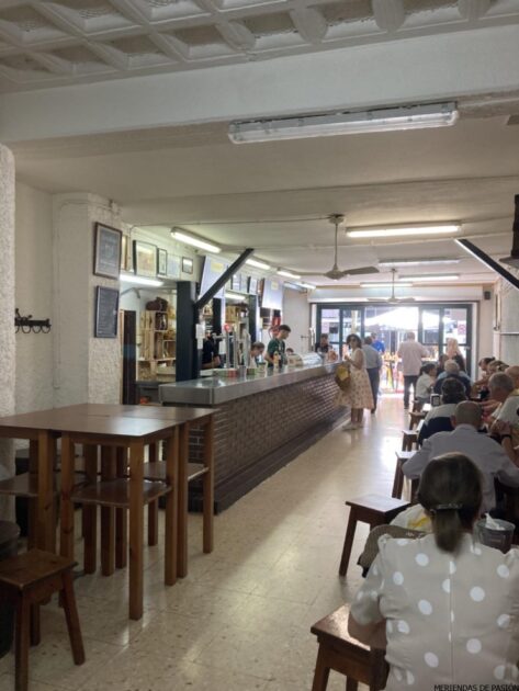 Interior de una animada cafetería con clientes en el mostrador y sentados en las mesas.