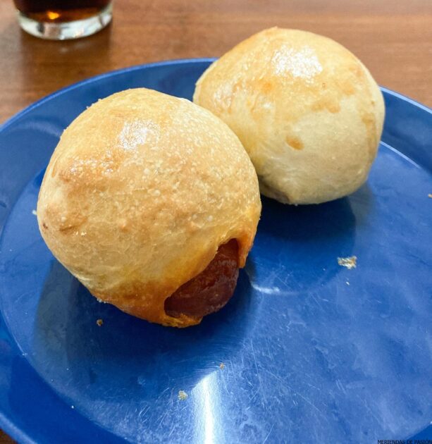 Dos bollos recién horneados en un plato azul con un vaso de bebida al fondo.