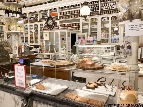 Interior de una panadería tradicional con una variedad de pasteles expuestos bajo cúpulas de cristal y un fondo de azulejos decorativos.