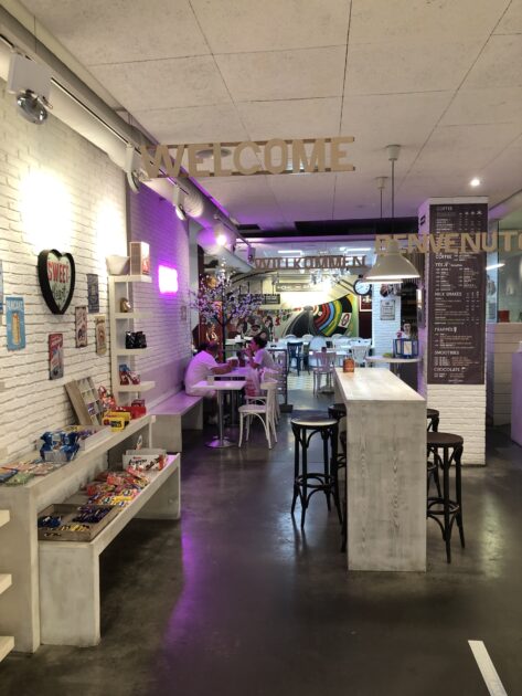 Un interior de cafetería acogedor y colorido con decoraciones eclécticas y un cartel de bienvenida en lo alto.