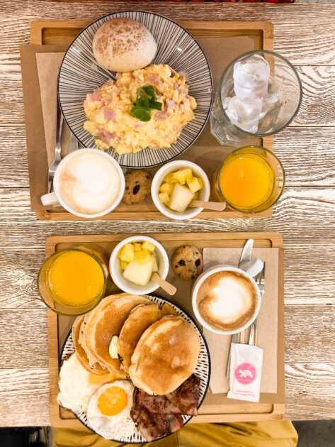 Una bandeja de desayuno con huevos revueltos, panqueques, huevos fritos, capuchinos, jugo de naranja y guarniciones sobre una mesa de madera.