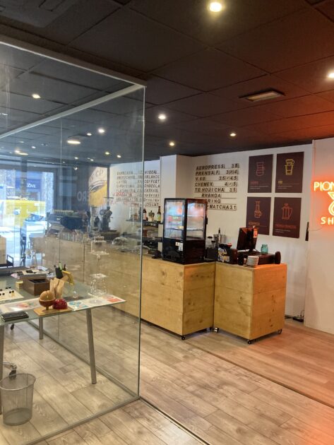Vista interior de una cafetería informal con carteles promocionales, un mostrador de vidrio que muestra productos y áreas para sentarse visibles a través de paredes de vidrio.