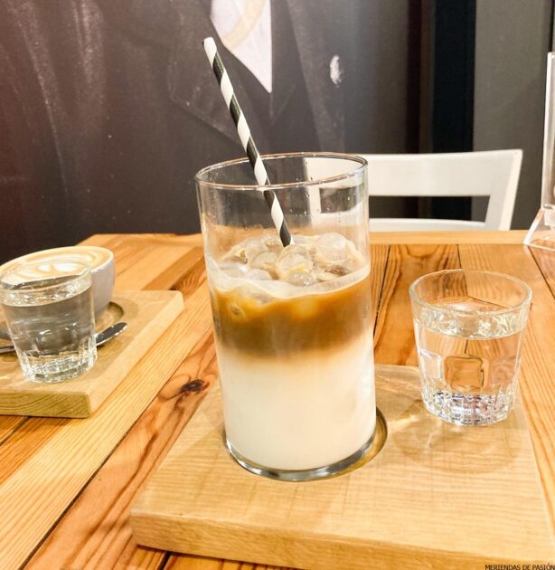 Un vaso de café con leche helado con una pajita negra sobre una tabla de servir de madera, acompañado de un vaso pequeño de agua.