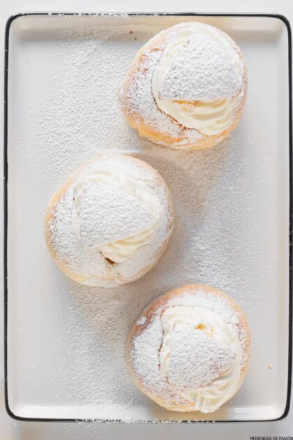 Tres pasteles rellenos de crema espolvoreados con azúcar en polvo en una bandeja para hornear.