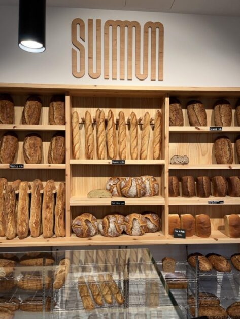 Una variedad de panes en estantes de madera en una panadería con el nombre "SUMMUM" en la pared de arriba.