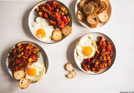 Tres platos con huevos fritos, pisto y rebanadas de pan tostado dispuestos sobre una superficie de madera clara.