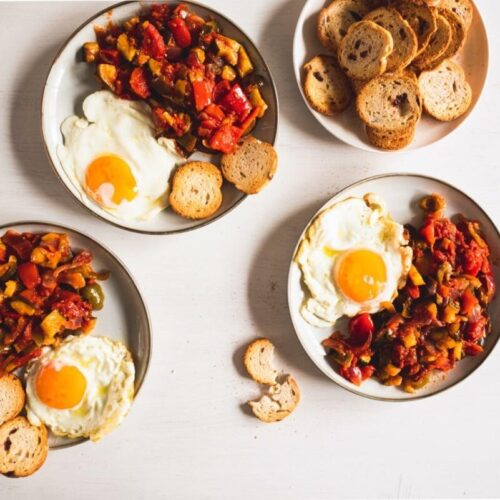 Tres platos con huevos fritos, pisto y rebanadas de pan tostado dispuestos sobre una superficie de madera clara.