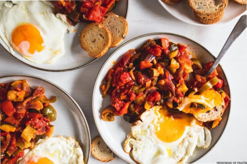 Un desayuno que incluye huevos fritos, tostadas y un plato de tomates y verduras salteados sobre una mesa blanca.
