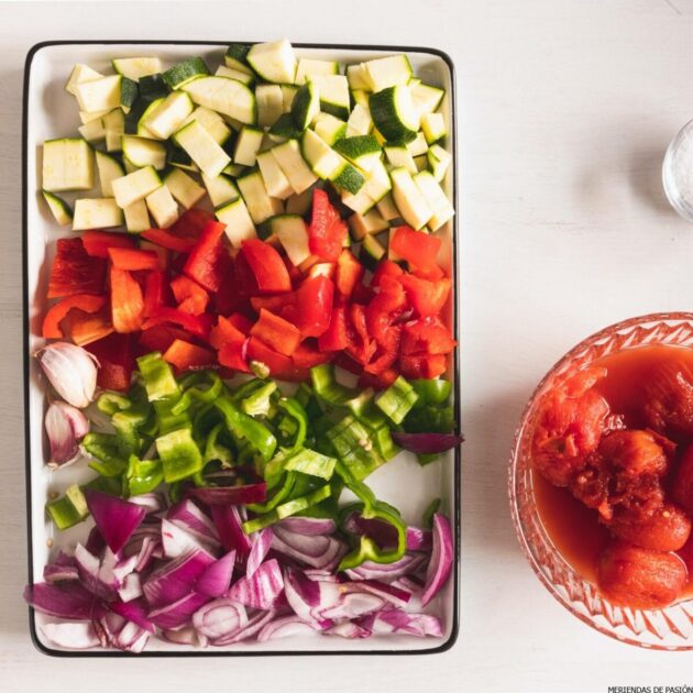 Chopped vegetables, including zucchinis, red peppers, green peppers, red onions, and garlic, on a cutting board, next to a plate of whole tomatoes.