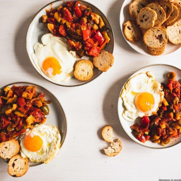 Three plates with ratatouille and fried eggs, accompanied by slices of toasted bread, arranged on a light-colored table.
