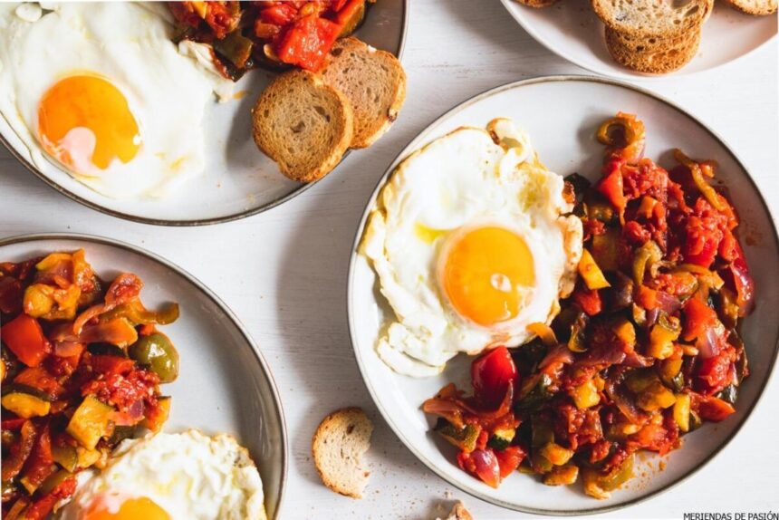 Three plates with fried eggs and ratatouille, accompanied by slices of toasted bread, served on a white table.