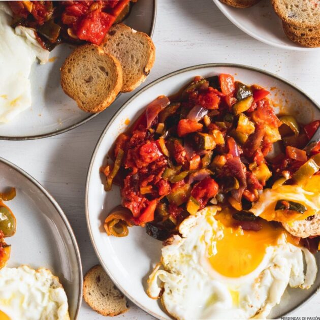 Plates of fried eggs with sautéed vegetables and slices of toasted bread served on a white table.