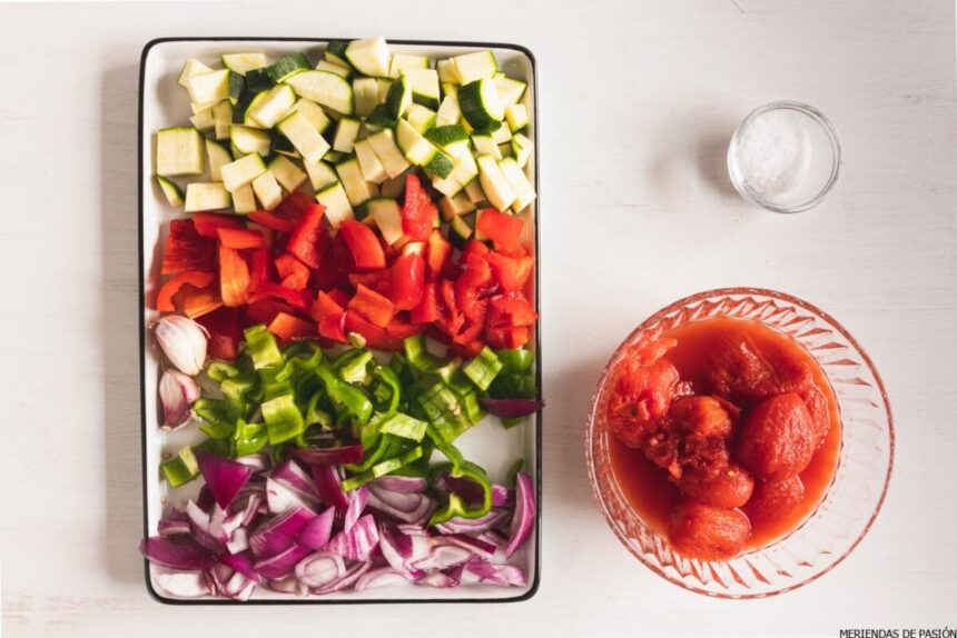 A tray of chopped vegetables including zucchinis, red peppers, green peppers, and purple onions, next to a plate of whole peeled tomatoes. enteros pelados.