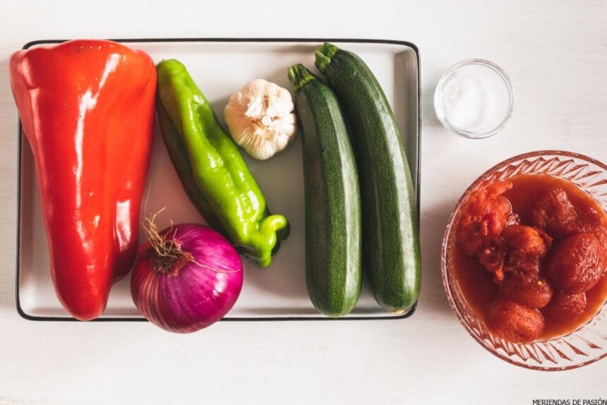 Verduras frescas variadas que incluyen pimiento rojo, chile verde, calabacín, cebolla morada, ajo y un plato de tomates en una bandeja.