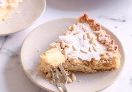 Un trozo de pastel cubierto de migas con un tenedor sobre un plato blanco. El pastel se adorna con azúcar en polvo y piñones, con porciones de pastel adicionales al fondo.