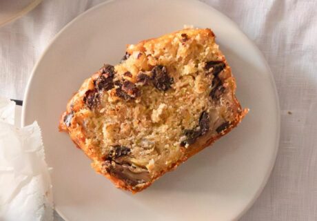 Una rebanada de pastel de nueces y chispas de chocolate en un plato blanco con un tenedor al lado sobre una mesa con un mantel blanco.