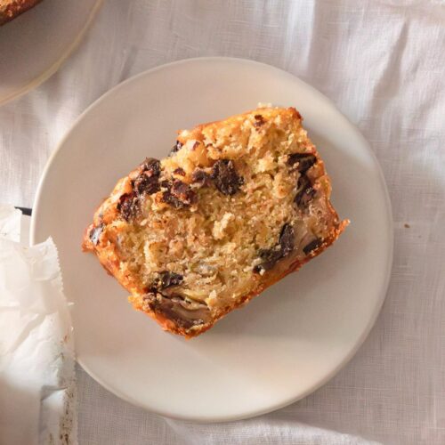 Una rebanada de pastel de nueces y chispas de chocolate en un plato blanco con un tenedor al lado sobre una mesa con un mantel blanco.