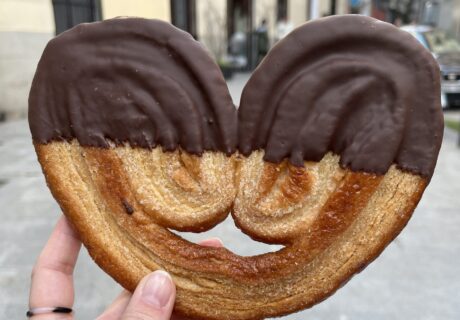 Una mano sosteniendo una palmera de hojaldre en forma de corazón, medio sumergido en chocolate, en una calle de la ciudad.