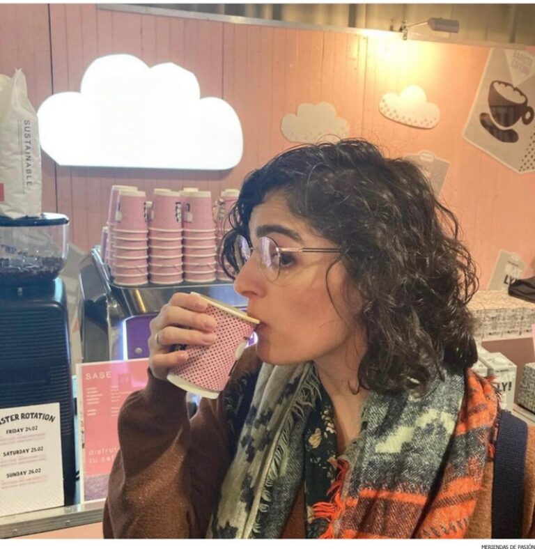 Una mujer con cabello rizado y anteojos bebe de un vaso de papel rosa en el mostrador de una cafetería decorado con recortes de nubes y vasos rosados apilados.
