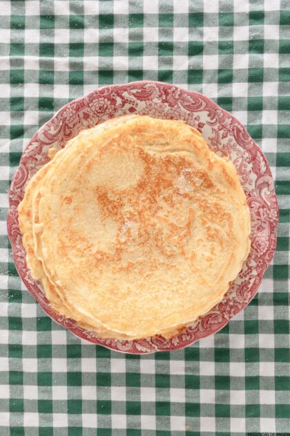 Una pila de crepes simples sobre un plato con estampado rojo y blanco reposa sobre un mantel a cuadros verdes y blancos.