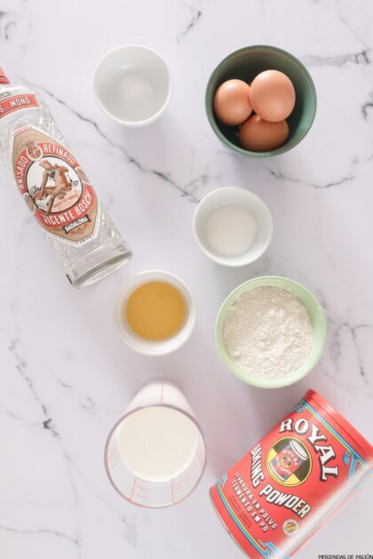 Ingredientes para hornear expuestos sobre una superficie de mármol: huevos, harina, levadura en polvo, azúcar, sal, extracto de vainilla, leche y una botella de alcohol.