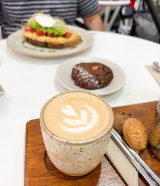 Una taza de café con leche con arte latte, galletas, una galleta de chocolate y un plato de tostadas con aguacate y verduras sobre una mesa blanca.