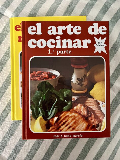 Dos libros de cocina españoles, uno titulado "El arte de cocinar 1.ª parte" de María Luisa García con imágenes de comida en la portada, están apilados sobre una superficie rayada.