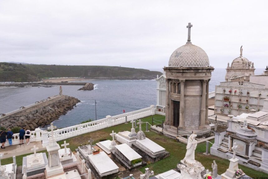 Un cementerio costero con varias tumbas, estatuas y mausoleos domina el mar bajo un cielo nublado, con gente caminando por un muelle cercano.