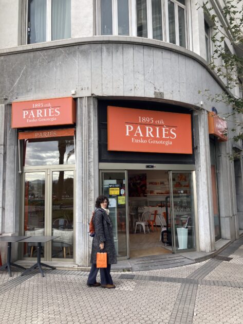 Una persona está de pie frente a una pastelería Paries con letreros naranjas, sosteniendo una bolsa de la compra también naranja. El local cuenta con grandes ventanales y una fachada curva de piedra.