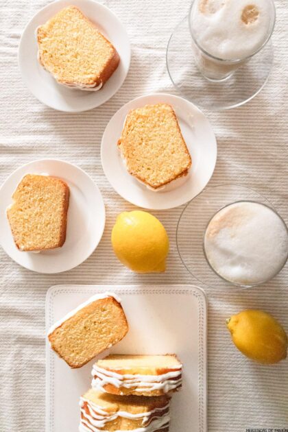 Rebanadas de pastel de limón en platos blancos, dos limones enteros y dos vasos de café espumoso dispuestos sobre un mantel blanco.