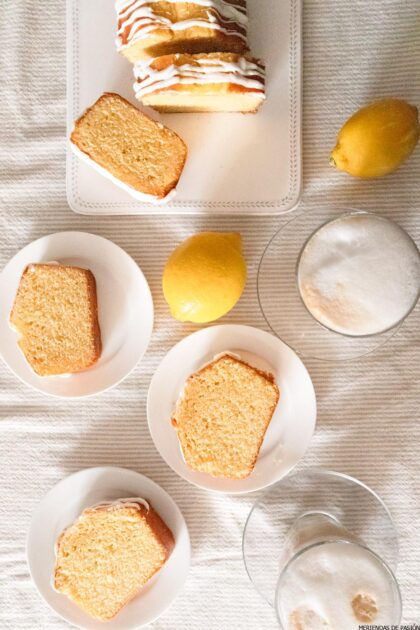 Pastel de limón en rodajas sobre platos y una bandeja para servir, con limones enteros y tazas de café espumoso sobre un mantel blanco.