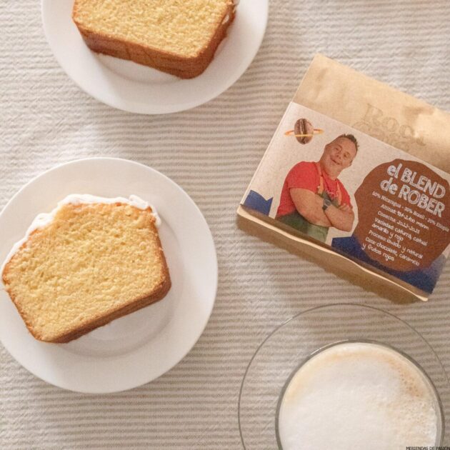 Dos rebanadas de bizcocho en platos blancos, una taza de café con espuma y un paquete de café etiquetado "el BLEND de ROBER" dispuestos sobre un mantel de rayas claras.