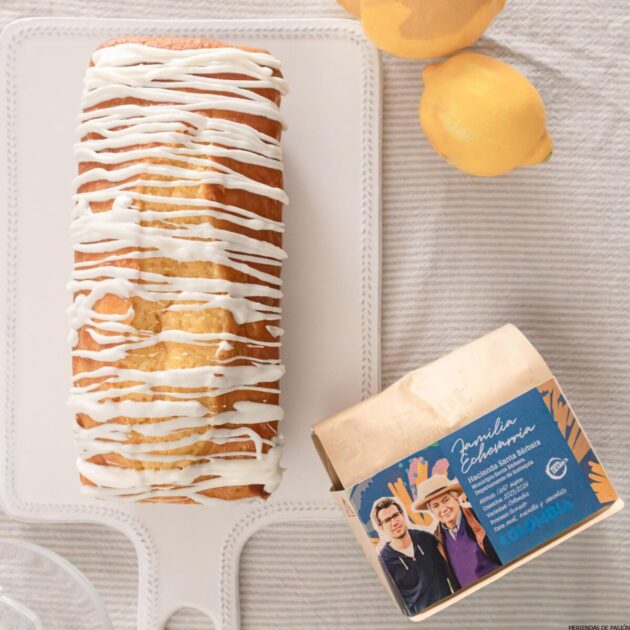Un trozo de pastel de limón con glaseado blanco sobre una bandeja blanca, junto a tres limones y una caja de café colombiano sobre una superficie de color claro.
