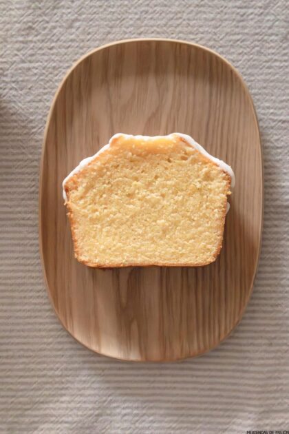 Se coloca una rebanada de bizcocho sencillo con un glaseado ligero sobre un plato de madera ovalado, dispuesto sobre un mantel texturizado de color claro.