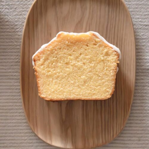 Se coloca una rebanada de bizcocho sencillo con un glaseado ligero sobre un plato de madera ovalado, dispuesto sobre un mantel texturizado de color claro.