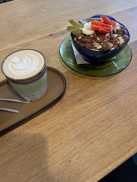Una taza de café con leche con arte latte se encuentra en una bandeja al lado de un tazón de granola cubierto con frutas en rodajas, colocado sobre una mesa de madera.