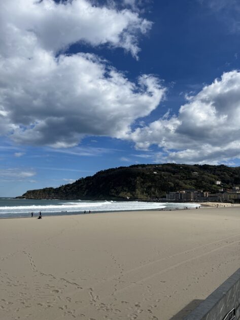 Amplia playa de arena con algunas personas paseando, olas que se acercan a la orilla, colina verde al fondo y cielo azul parcialmente nublado en lo alto.