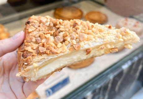 Una mano sostiene una rebanada de masa de hojaldre rellena de crema y cubierta con nueces picadas, frente a la vitrina de una panadería.