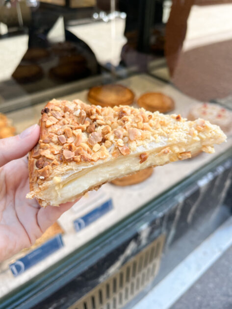 Una mano sostiene una rebanada de masa de hojaldre rellena de crema y cubierta con nueces picadas, frente a la vitrina de una panadería.