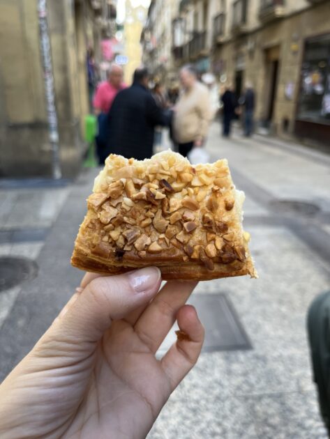Una mano sostiene un trozo de pastel cubierto con nueces picadas en una calle de la ciudad; en el fondo aparecen peatones y edificios borrosos.