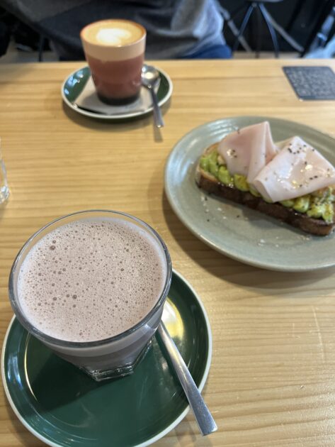 Un vaso de chocolate caliente, un plato con tostadas de aguacate coronadas con jamón y una taza de café en una mesa de madera.