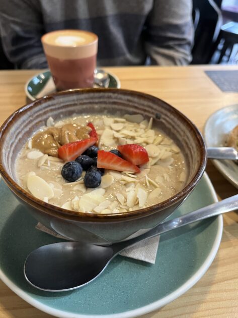 Un tazón de avena cubierto con mantequilla de maní, almendras fileteadas, arándanos y fresas reposa sobre un platillo con una cuchara; una taza de café y una persona en el fondo.