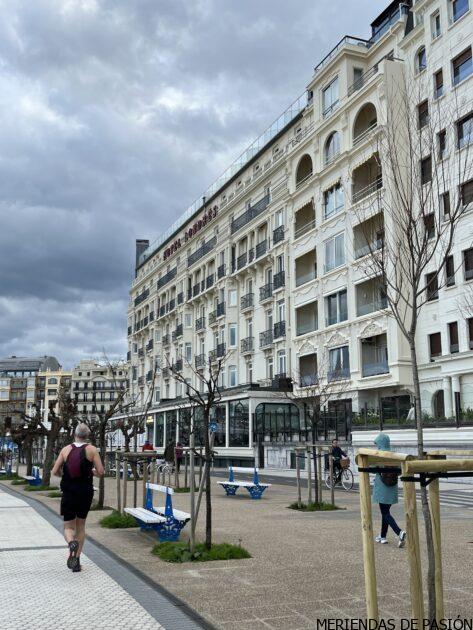 Una persona hace footing por un paseo bordeado de árboles y bancos, con un gran edificio hotelero de varias plantas al fondo, bajo un cielo nublado.