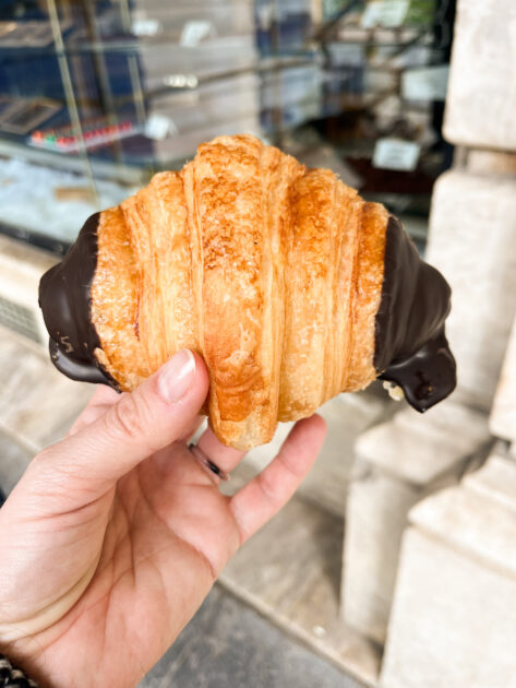 Una mano sosteniendo un croissant con ambos extremos sumergidos en chocolate, fotografiada al aire libre frente al escaparate de una tienda.