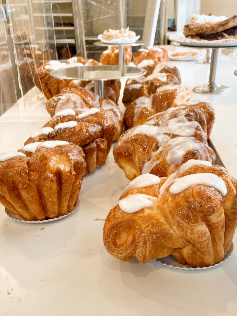 Una fila de pasteles con glaseado blanco encima, dispuestos sobre una encimera blanca en una panadería, con más productos horneados exhibidos en el fondo.
