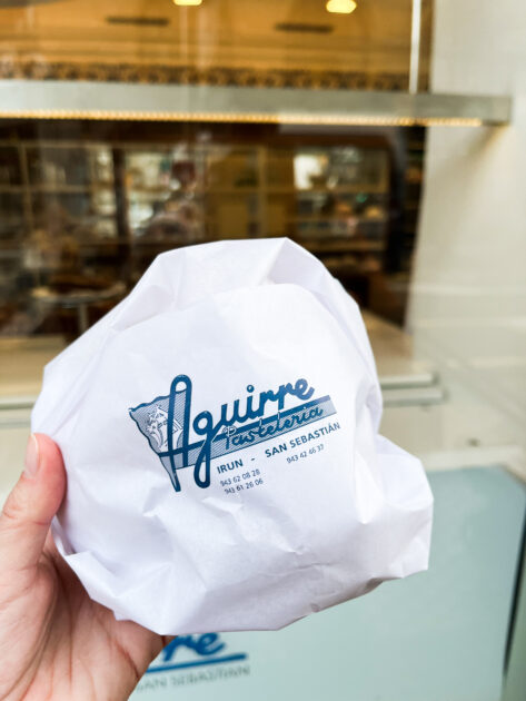 Una mano sostiene un artículo envuelto en papel blanco con el logotipo de Aguirre Pastelería, con direcciones de Irún y San Sebastián.