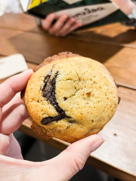 Una mano sostiene una galleta con chispas de chocolate con un remolino de chocolate visible; en el fondo, en una mesa de madera, hay una persona borrosa con un menú.